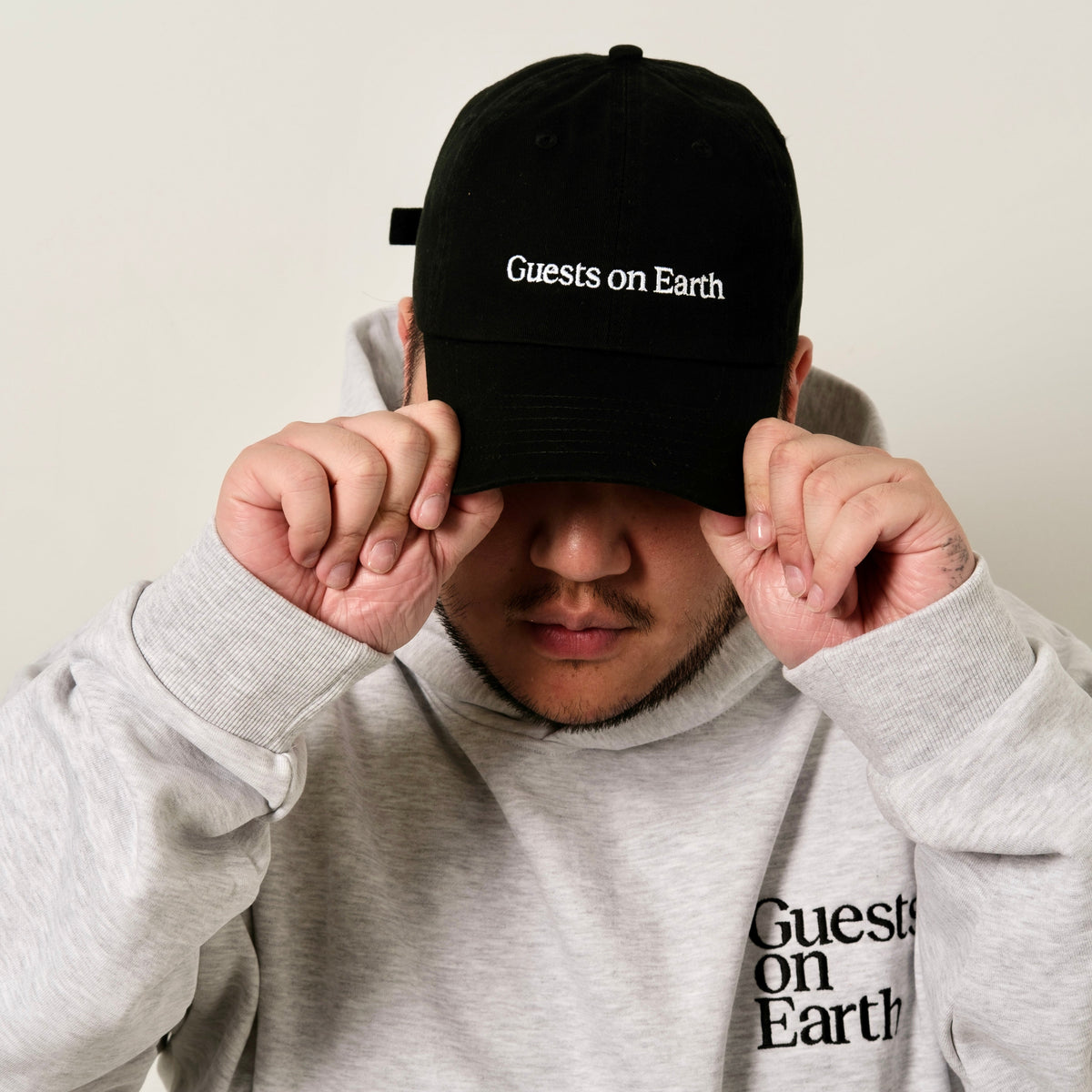 Guests on Earth black ambassador cap with white logo, worn by model adjusting the brim.