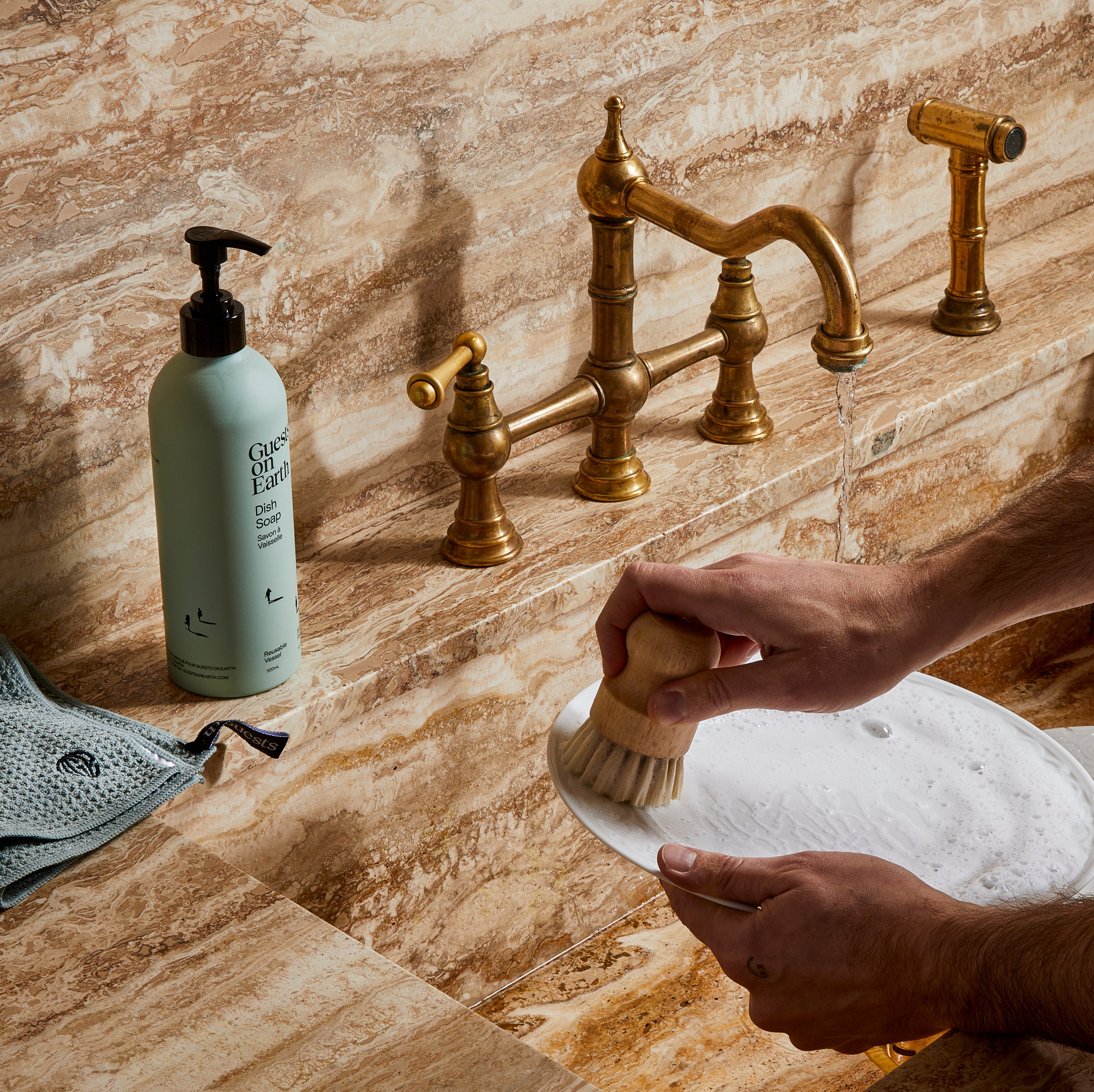 Guests on Earth Dish Soap beside brass tap as hands scrub a soapy plate with a wooden brush at the sink.