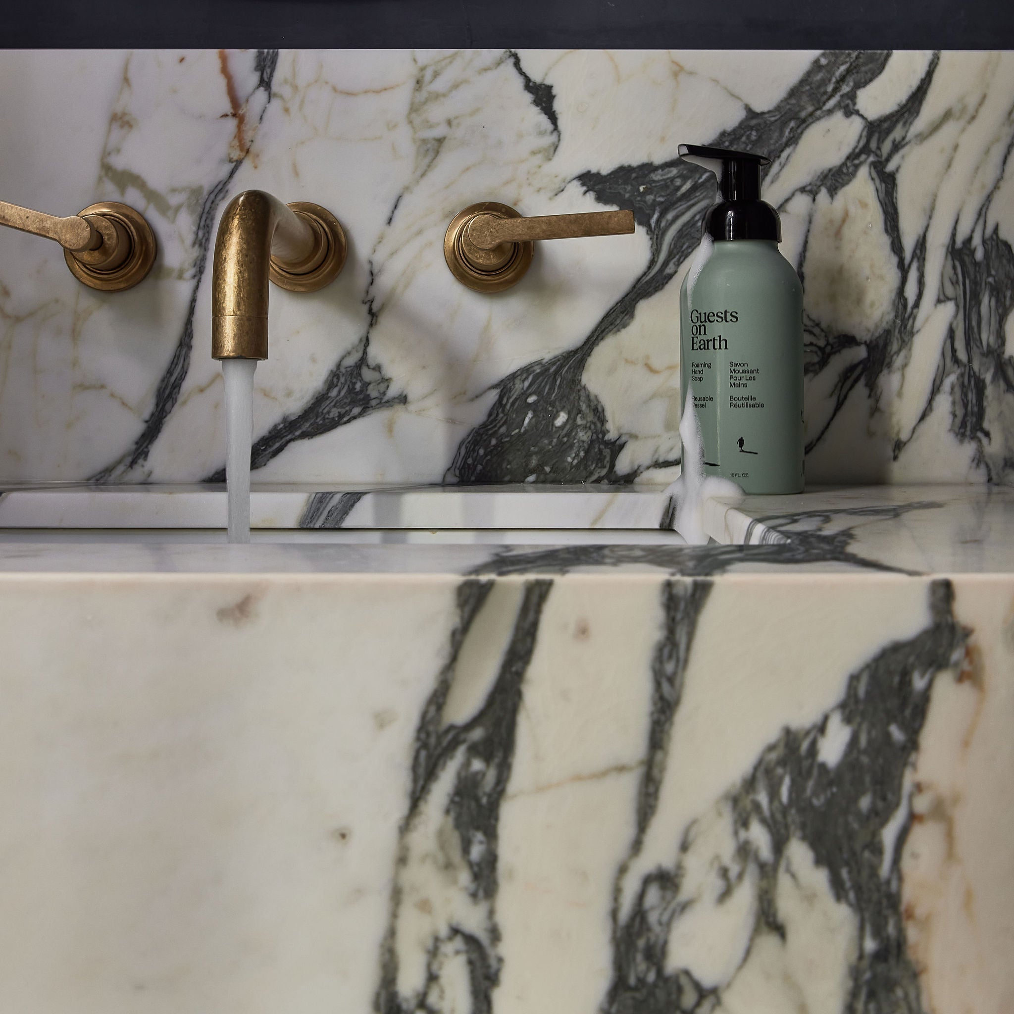 Guests on Earth Foaming Hand Soap on marble sink ledge beside brass tap with running water and a drip of foam
