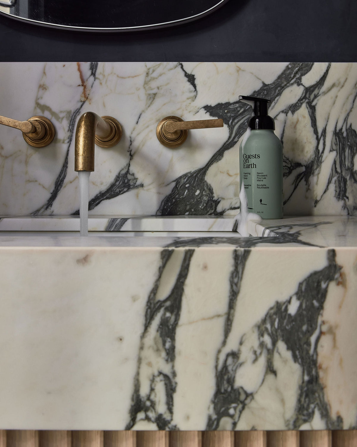 Guests on Earth Foaming Hand Soap on marble sink ledge beside brass tap with running water and a drip of foam